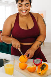 Happy healthy female cutting fruits in kitchen. Plus size woman in workout wear making juice at home. - JLPSF03723