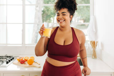 Plus size female with glass of fruit juice while standing at kitchen. Pretty woman in workout wear smiling and holding glass of fruit juice at home. - JLPSF03728
