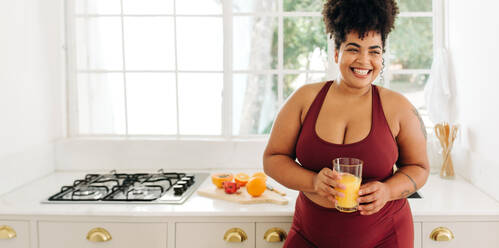 Wide angle shot of plus size female smiling while holding glass of fruit juice at home. Happy woman with fruit juice standing at kitchen. - JLPSF03729