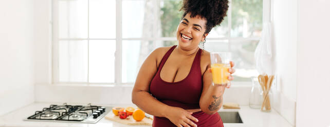 Wide angle shot of fitness woman smiling while standing with glass of fruit juice. Plus size female having fruit juice at home. - JLPSF03730