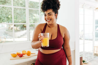 Beautiful female smiling and holding glass of fruit juice in kitchen. Pretty woman in sportswear with fresh fruit juice after workout at home. - JLPSF03746