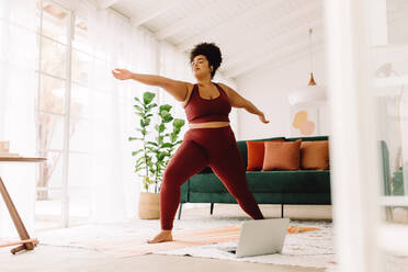 Full length of body positive woman exercising at home with laptop on floor. Pretty female in fitness wear doing workout at home. - JLPSF03756