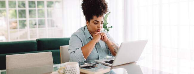 Frau sitzt auf dem Tisch und schaut auf den Laptop. Frau sitzt bei der Einnahme im Wohnzimmer und arbeitet am Laptop-Computer zu Hause. - JLPSF03833