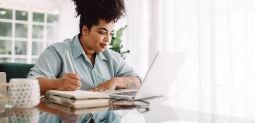 Geschäftsfrau arbeitet von zu Hause aus und schreibt Notizen, während sie auf den Laptop schaut. Selbstbewusste Frau sitzt am Schreibtisch, benutzt den Laptop und macht sich Notizen. - JLPSF03839