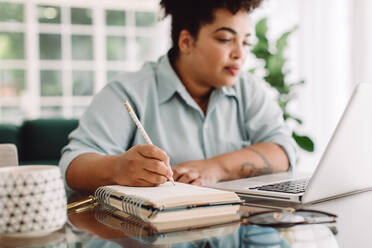 Eine Frau schreibt Notizen in ihr Tagebuch, während sie von zu Hause aus arbeitet. Eine Geschäftsfrau schaut auf ihren Laptop und macht sich Notizen in ihrem Tagebuch im Büro. - JLPSF03844