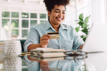 Attractive female sitting at desk smiling while holding credit card in hand. Beautiful woman with credit card and laptop doing online shopping. - JLPSF03847