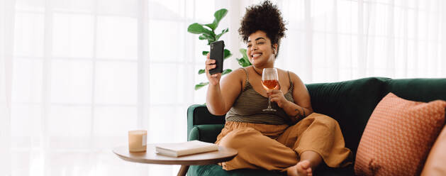 Wide angle shot of attractive female sitting on sofa holding a glass of wine and making video call on mobile phone. Beautiful woman sitting on couch having virtual happy hour on video call. - JLPSF03861