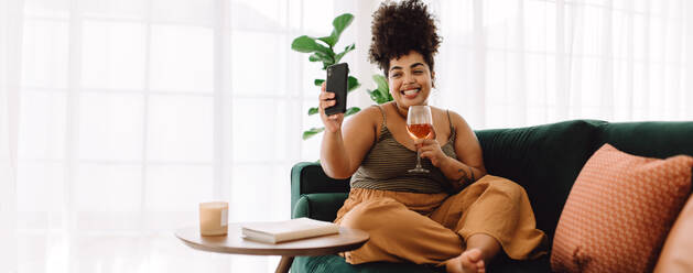 Beautiful woman sitting on sofa holding a glass of wine and making video call on mobile phone. Female sitting on couch having virtual happy hour on video call. - JLPSF03862