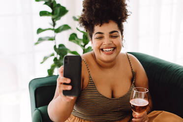 Schöne Frau sitzt lächelnd auf dem Sofa und macht ein Selfie mit dem Handy, während sie ein Glas Wein in der Hand hält. Frau macht Selfie mit einem Glas Wein. - JLPSF03863
