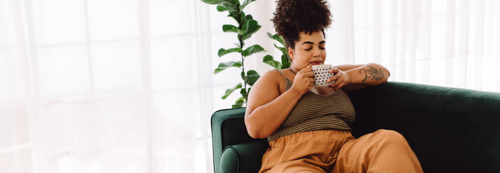 Wide angle shot of beautiful female having coffee at home. Woman sitting on couch and drinking coffee. - JLPSF03869