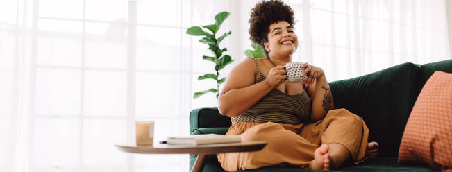 Happy female holding coffee mug while sitting on couch at home. Smiling woman sitting on sofa having coffee. - JLPSF03872