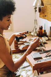Eine Künstlerin fertigt in ihrem Atelier mit einem Brenner und Werkzeugen Schmuck an. Eine junge Juwelierin fertigt an ihrem Schreibtisch in der Werkstatt handgefertigte Ornamente an. - JLPSF03909
