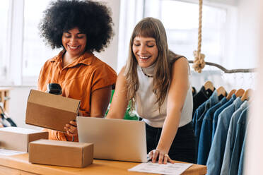 Glückliche Geschäftsfrauen, die in einem Secondhand-Laden einen Laptop benutzen, während sie Pakete für die Lieferung vorbereiten. Zwei Kleinunternehmerinnen, die eine Lieferung planen. Unternehmerinnen, die ein Online-Kleidungsgeschäft betreiben. - JLPSF03929