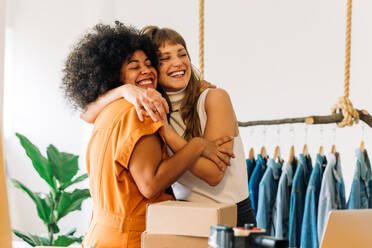 Glückliche Online-Shop-Besitzerinnen, die sich in ihrem Secondhand-Laden umarmen. Zwei fröhliche Geschäftsfrauen, die ihren Erfolg als Team feiern. Zwei Unternehmerinnen, die ein kleines E-Commerce-Unternehmen betreiben. - JLPSF03932