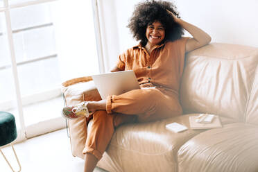 Fröhliche schwarze Geschäftsfrau, die in die Kamera lächelt, während sie mit einem Laptop in einer Bürolobby sitzt. Glückliche Unternehmerin, die allein an einem modernen Arbeitsplatz arbeitet. - JLPSF03944