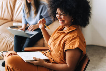 Schwarze Geschäftsfrau lächelt fröhlich, während sie mit einem Notebook in einer Bürohalle sitzt. Glückliche junge Geschäftsfrau, die mit ihrem Kollegen in einem Büro arbeitet. - JLPSF03962