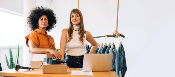 Zwei Inhaberinnen eines Bekleidungsgeschäfts schauen in ihrem Laden in die Kamera. Geschäftsfrauen bereiten in ihrem Secondhand-Laden Pakete für den Versand vor. Unternehmerinnen, die ein kleines E-Commerce-Geschäft betreiben. - JLPSF03978