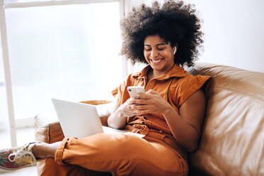 Cheerful young businesswoman using a smartphone while working in an office lobby. Happy young businesswoman reading a text message while sitting on a couch in a modern workplace. - JLPSF04002