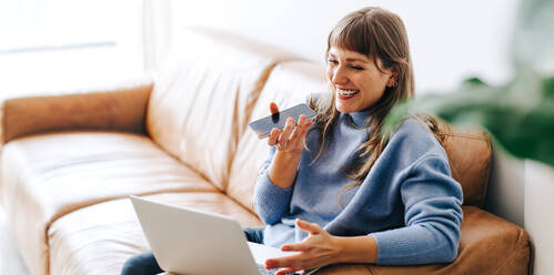 Happy young businesswoman having a phone call discussion while sitting on a couch. Cheerful female entrepreneur speaking on the phone while working with a laptop in a modern office. - JLPSF04013