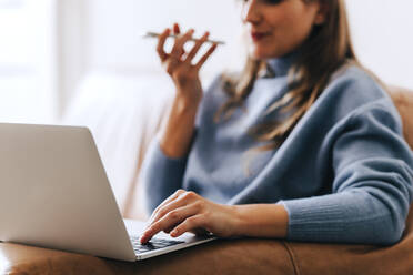 Geschäftsfrau, die einen Anruf tätigt, während sie an einem Laptop an einem modernen Arbeitsplatz arbeitet. Junge Frau sitzt auf einer Couch in einer Büro-Lobby. - JLPSF04014