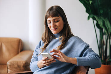 Businesswoman using a mobile phone while sitting in an office lobby. Young female entrepreneur reading a text message while working in a modern workplace. - JLPSF04038