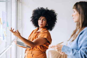 Junge Geschäftsfrauen beim Brainstorming während einer Besprechung. Zwei junge Geschäftsfrauen diskutieren ihre Geschäftsideen mit Hilfe von Haftnotizen. Kreative Unternehmerinnen arbeiten als Team in einem modernen Büro. - JLPSF04042