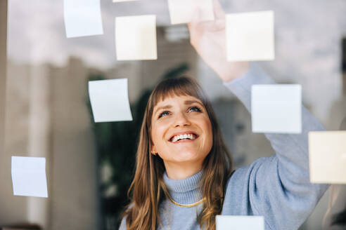 Happy young businesswoman sticking adhesive notes to a glass wall in a modern office. Cheerful female entrepreneur laying out her ideas while working on a new creative strategy. - JLPSF04046