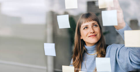 Junge Unternehmerin, die in einem modernen Büro Haftnotizen an eine Glaswand klebt. Glückliche junge Geschäftsfrau, die bei der Arbeit an einer neuen kreativen Strategie ihre Ideen festhält. - JLPSF04047