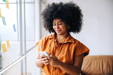 Cheerful black businesswoman using a smartphone in a creative office. Young female entrepreneur reading a text message while standing next to a glass wall with sticky notes. - JLPSF04056