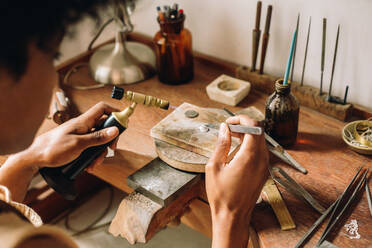 Kunsthandwerker bei der Herstellung von Schmuckdesign mit Flammenbrenner und Werkzeug in der Werkstatt. Juwelierin bei der Arbeit am Schreibtisch mit traditionellen Werkzeugen in ihrem Atelier. - JLPSF04069