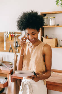 Accessoires-Unternehmerin bespricht Details einer Schmuckbestellung am Telefon in einem Atelier. Frau spricht am Handy mit ihrem Buch in einer Schmuckwerkstatt. - JLPSF04095