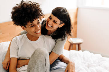 Lesbian couple laughing cheerfully at home. Affectionate young woman embracing her girlfriend warmly while sitting on their bed at home. Happy young LGBTQ+ couple bonding in their bedroom. - JLPSF04169