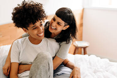 Lesbisches Paar, das zu Hause fröhlich lacht. Liebevolle junge Frau, die ihre Freundin herzlich umarmt, während sie zu Hause auf dem Bett sitzt. Glückliches junges LGBTQ+ Paar, das sich in ihrem Schlafzimmer zusammenfindet. - JLPSF04169