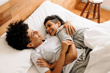 Happy couple cuddling on their bed at home. Romantic young LGBTQ+ couple embracing each other while lying down together in their bedroom. Smiling lesbian couple bonding fondly at home. - JLPSF04173