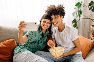 Lächelndes lesbisches Paar, das ein Selfie mit Popcorn zu Hause macht. Glückliches lesbisches Paar, das für ein Selfie in seinem Wohnzimmer posiert. Junges LGBTQ+ Paar, das Zeit miteinander verbringt. - JLPSF04181