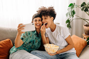 Fröhliches lesbisches Paar, das ein Selfie mit Popcorn zu Hause macht. Glückliches lesbisches Paar, das für ein Selfie in seinem Wohnzimmer posiert. Junges LGBTQ+ Paar, das Zeit miteinander verbringt. - JLPSF04182