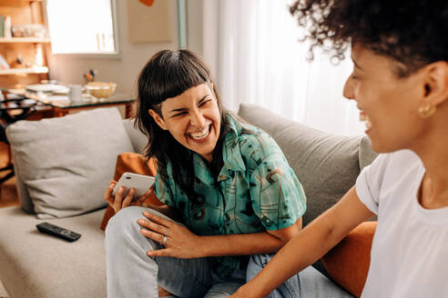 Woman laughing with her wife at home. Happy young lesbian woman laughing cheerfully while sitting with her wife in their living room at home. Young LGBTQ+ couple bonding at home. - JLPSF04183