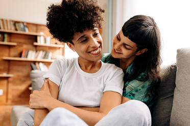 Happy lesbian lovers embracing each other at home. Two romantic female lovers smiling cheerfully while sitting together in their living room at home. Young LGBTQ+ couple bonding fondly at home. - JLPSF04187