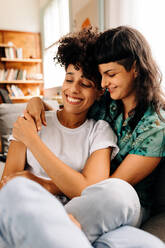 Young lesbian couple cuddling each other. Affectionate young lesbian couple smiling cheerfully while sitting together in their living room. Two romantic female lovers bonding fondly at home. - JLPSF04192