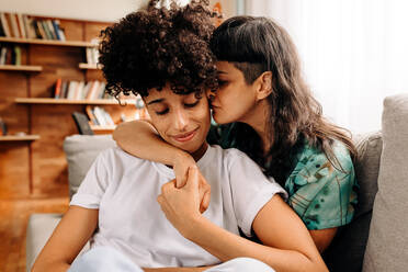 Affectionate young lesbian couple bonding at home. Young lesbian couple embracing each other while sitting in their living room. Two romantic female lovers spending quality time together at home. - JLPSF04193