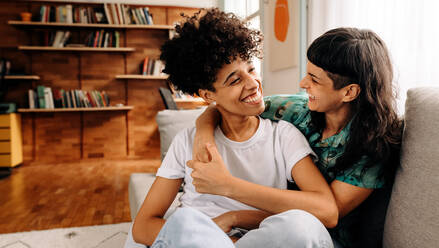 Cheerful female lovers laughing together. Happy young lesbian couple sharing a laugh while sitting together in their living room. Young lesbian couple spending quality time together. - JLPSF04194