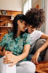 Carefree lesbian couple spending quality time together. Happy married lesbian couple smiling cheerfully while sitting together in their living room. Romantic young LGBTQ+ couple bonding at home. - JLPSF04198