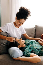 Happy female lovers relaxing together at home. Happy young woman smiling at her girlfriend while laying her head on her lap. Young lesbian couple bonding fondly in their living room at home. - JLPSF04199