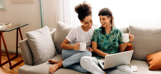 Online-Shopping und Chillen. Glückliches junges lesbisches Paar genießt das Online-Shopping, während es sich in seinem Wohnzimmer zu Hause entspannt. Lächelnde weibliche Liebhaber, die eine schöne Zeit miteinander verbringen. - JLPSF04202