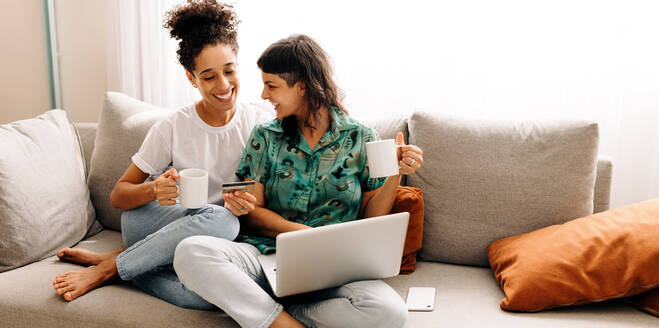 Sie genießen die Online-Wochenendrabatte. Ein glückliches junges lesbisches Paar kauft online ein und entspannt sich dabei in ihrem Wohnzimmer zu Hause. Lächelnde weibliche Liebhaber, die eine schöne Zeit miteinander verbringen. - JLPSF04203