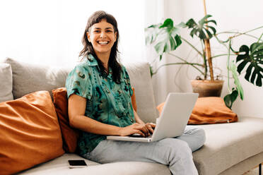 Glückliche junge Frau, die ihren persönlichen Blog zu Hause aktualisiert. Junge Frau, die in die Kamera lächelt, während sie einen Laptop auf einer Couch benutzt. Junge Frau, die sich allein in ihrem Wohnzimmer zu Hause entspannt. - JLPSF04207