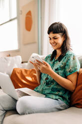 Young woman reading a text message on her smartphone. Happy young woman smiling cheerfully while holding a smartphone on her couch. Young woman sitting with a laptop in her living room. - JLPSF04214