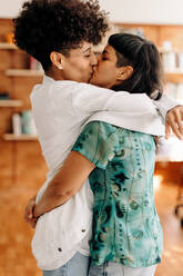 Cute lesbian couple kissing indoors. Two romantic female lovers embracing each other affectionately. Young LGBTQ+ couple being romantic while standing indoors at home. - JLPSF04215