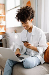 Fröhliche junge Frau beim Online-Einkauf. Junge Frau lächelt, während sie mit ihrer Kreditkarte auf ihrem Laptop einkauft. Frau sitzt allein auf einer Couch zu Hause. - JLPSF04217