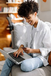 Frau macht einen Online-Einkauf auf ihrem Laptop. Glückliche junge Frau lächelt fröhlich beim Online-Shopping zu Hause. Junge Frau sitzt allein auf einer Couch in ihrem Wohnzimmer. - JLPSF04218